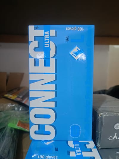 The image shows a blue box of "CONNECT ULTRA" disposable gloves. The box is standing upright on a shelf, partially obscuring another gray box. The blue box has large white lettering that spells out "CONNECT" vertically along its side. The top of the blue box shows text indicating "100 GLOVES" and "SIZE L" along with a stylized blue handprint icon. The gray box appears to have "y" and "100 pairs" written on it. The background shows additional boxes and shelves. The scene is indoors, likely a storage area or a retail environment. The image does not provide any information about the location in Berdiansk, Ukraine.