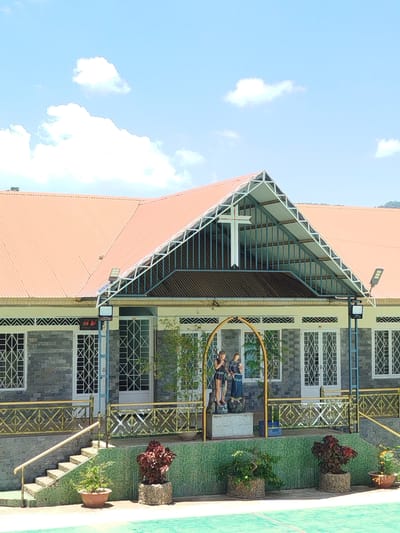 The image shows a building with a prominent orange roof and a large white cross on its facade, suggesting it is a church or religious building. The building's exterior walls are clad in gray stone. A porch area features a yellow metal railing and a set of stairs leading up to the entrance. In front of the building, there are several potted plants. To the left of the entrance, two statues of individuals are visible behind a decorative arched structure. The sky is blue with scattered white clouds. The building appears to be located in a relatively rural or suburban setting, possibly K'Rèn, Vietnam, given the context. There are no people actively engaged in any discernable actions within the scene.