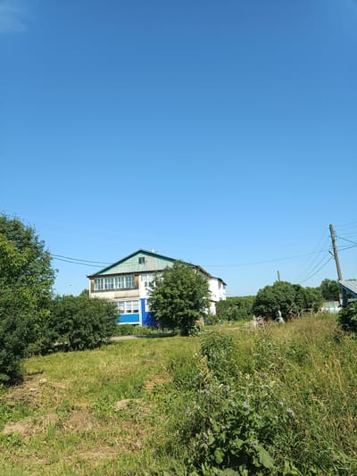 The image depicts a two-story building with a green, sloped roof, possibly a house, situated in a rural or semi-rural area. The building has white and blue sections, with a balcony on the upper floor featuring white framed windows. It is surrounded by lush green trees and dense vegetation, including tall grasses and flowering plants. A wooden utility pole with electrical wires is visible to the right of the building. The sky is a clear, bright blue. The scene appears to be a daytime shot, under sunny conditions, likely in a village or dacha settlement. The specific location in the city of Посад, Russia, cannot be determined from the visual information alone.