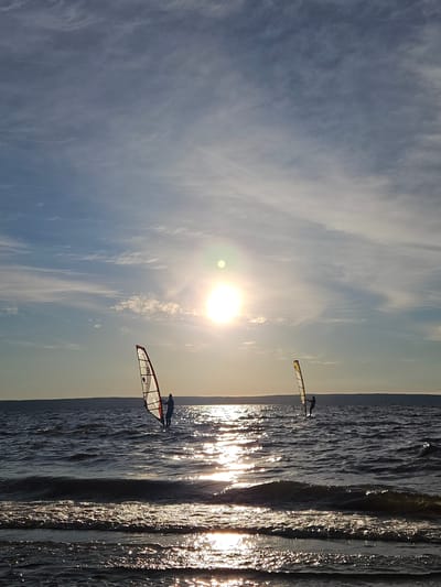 In Leninsky, Russia, two individuals are windsurfing on a body of water under a bright, partially obscured sun. The sky is a mix of blue and wispy white clouds. The sun, a bright orb, is positioned high in the sky, casting a shimmering reflection across the water's surface. The water itself is choppy, with visible waves and whitecaps. The shoreline in the foreground is dark and wet, with small waves lapping at it. The two windsurfers are silhouetted against the bright sky and sun. The windsurfer on the left has a sail that appears to be red and white with the number '9' visible. The windsurfer on the right has a predominantly yellow sail. Both are standing on boards and holding onto their respective sails. In the distance, a dark, flat horizon line suggests land. The overall scene depicts an active, outdoor recreation scenario during daylight hours.