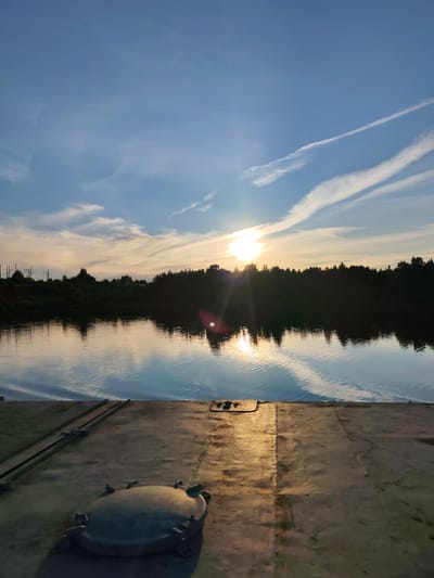 This media file captures a serene sunset scene over a body of water, likely a river or lake, in Tarkanovo, Russia. The foreground is dominated by a weathered, metallic surface, possibly a deck or pier, with a large, round hatch cover in the lower left corner. The metallic surface shows signs of age and use, with some areas reflecting the golden light of the sunset.
Beyond the metallic foreground, the calm water mirrors the sky and the tree line on the opposite bank. The water is rippled, creating a textured surface that reflects the light. The opposite bank is lined with a dense, dark silhouette of trees, creating a stark contrast with the bright sky.
The sky is a prominent feature, displaying a gradient of colors from deep blue at the top to softer hues of yellow and orange near the horizon. Wispy clouds are scattered across the sky, catching the sunlight and adding texture. The sun itself is partially obscured by clouds and trees, casting a bright, diffused light and creating lens flare effects. The reflection of the sun is visible on the water.
The overall mood is peaceful and tranquil, suggesting a quiet moment at the end of the day. The time of day is clearly sunset, as indicated by the low angle of the sun and the warm colors in the sky. There is no visible text in the image. The location cues, such as the type of vegetation and the presence of what appear to be power lines in the distance on the left bank, are consistent with a rural or semi-rural setting in Russia. No people are visible in the image.