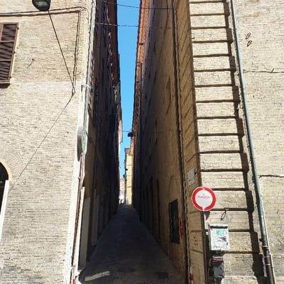 This image depicts Vicolo della Lupa, a narrow pedestrian alleyway in the city of Ancona, Italy. The alley is framed by two tall, multi-story buildings constructed of light-colored brick, which almost meet overhead, allowing only a sliver of clear blue sky to be visible.

The building on the left features a dark brown wooden shutter on an upper window and a high-mounted dark lamp fixture with associated wiring. An arched doorway is partially visible at the lower left. The building on the right displays a vertical metal conduit pipe and a series of prominent horizontal brick projections that cast distinct, ladder-like shadows onto its sunlit surface. Mounted on this wall are a red and white circular 'No Entry' sign and two utility boxes, the upper one covered in graffiti. A small white sign below the "No Entry" sign identifies the street as 'VIA DELLA LUPA'.

The cobbled ground of the alley slopes gently downward into the distance, appearing dark due to heavy shadow, contrasting with the bright sunlit right wall. A lone human figure is visible walking away in the extreme distance, at the far end of the alley where it widens slightly. The scene is illuminated by strong sunlight, casting sharp shadows.
