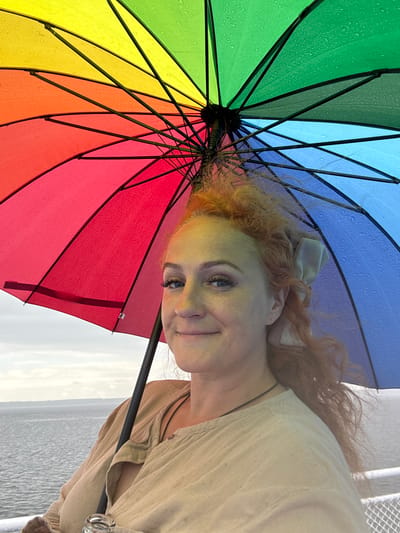 A medium shot captures a woman with red, curly hair under a vibrant, multi-colored umbrella as she looks towards the camera with a slight smile. The umbrella, open and displaying segments of red, orange, yellow, green, and blue, has visible raindrops on its surface, suggesting it is raining. The woman is wearing a light-colored, loose-fitting top. She has makeup on, including eyeliner and mascara. Her hair is partially tied back with a light-colored ribbon. The background shows a body of water, likely a sea or large lake, with a hazy sky above and a distant landmass on the horizon. A railing or edge of a boat is visible in the foreground, and a clear glass bottle with a metal cap is held by the woman's left hand, partially obscured. The overall setting appears to be outdoors, possibly on a ferry or boat, with a cool, overcast atmosphere. The location is identified as Белоус, Russia.