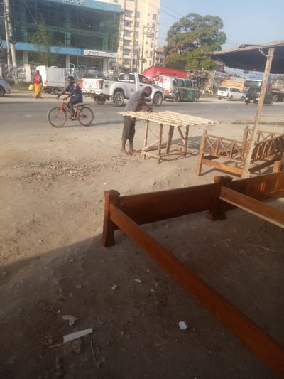The scene depicts a roadside carpentry operation and urban street activity in Ziwa La Ng'ombe ward, Kenya. In the immediate foreground, a dark reddish-brown wooden bed frame component lies horizontally on a dry, dirt ground littered with small debris. Shadows are cast across the lower right.
In the midground, a male individual, wearing a light-colored striped shirt and dark shorts, stands barefoot next to a simple wooden workbench. He is actively sanding or working on several light-colored wooden slats arranged horizontally on the workbench. To his right, several unfinished wooden furniture pieces, including benches with decorative lattice work, are displayed on the ground. To the left of the carpenter, a male cyclist wearing a dark blue shirt and dark shorts rides an orange bicycle on a paved road, moving from left to right across the frame.
The background reveals a bustling urban street with multiple lanes. Various vehicles are present, including a white Isuzu D-Max pickup truck, a multi-colored minivan (matatu), a red truck, and other cars, indicating active traffic. On the far left, a woman in a red top and yellow skirt carries a yellow bag while walking on a sidewalk. Beyond the road, multi-story buildings line the street; one building features a blue glass facade and "PRIME AUTO" signage, while another is a taller, beige apartment or office structure. A large green tree is visible above the buildings on the right side of the background. The sky is clear and light blue, indicating bright daylight.