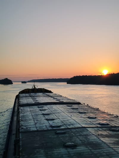 The image depicts a barge traversing a waterway at sunrise. The barge is large, gray, and flat, with a grid pattern of hatch covers across its deck. A yellow structure sits atop the barge towards the center of the frame. The water reflects the warm hues of the rising sun. The river is flanked by land on either side, with dense forests on the right and smaller islands on the left. Another vessel can be seen in the distance. The sky is clear and transitions from light blue to orange near the horizon. The location is unspecified and information about it being in Poteryaevo, Russia, is not provided within the image.