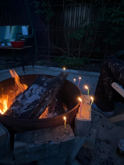The image depicts an outdoor scene at dusk, possibly in a backyard or garden area, illuminated by a bonfire and several lit candles. The bonfire is contained within a large, dark metal fire pit and contains burning logs. Several thin, colored candles are stuck into bricks or small concrete blocks arranged around the fire pit, some appearing to be drippy with wax. In the background, a dark fence with vertical slats is visible, partially obscured by leafy branches. To the left, a portion of a table with a red bowl and what appears to be a chair or bench with blue and white cushions can be seen. On the right side of the image, the lower half of a person's legs is visible, wearing dark patterned trousers and light blue shoes. A portion of a white object, possibly a chair or table leg, is also present. The ground appears to be a mix of gravel and dirt. A bottle with a Corona Extra label is visible in the lower right corner. The overall atmosphere suggests a casual gathering or celebration, possibly a birthday, given the presence of candles. There is no specific indicator that this scene is located in Вельяминово, Russia.