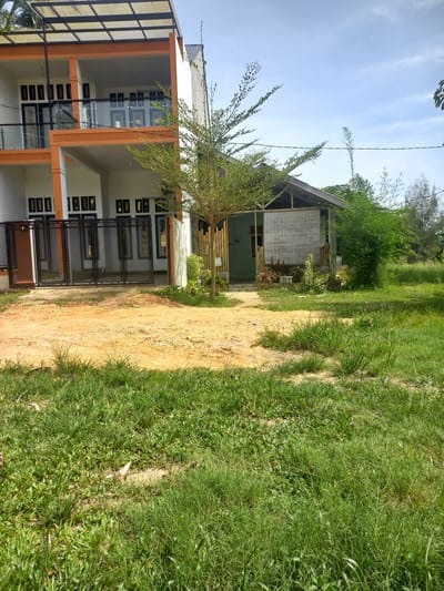 The image captures a scene in Gampongaceh, Indonesia, showcasing a two-story modern building with white walls and orange accents, featuring multiple balconies with glass railings and dark-framed windows. A dark metal gate is visible in front of the ground floor. Adjacent to the modern building, a smaller, more rustic structure with a wooden frame and a thatched or bamboo-covered wall stands under a gabled roof. Several trees and dense green vegetation surround the buildings, with a patch of sandy ground in the foreground partially covered by lush green grass. The sky is visible with some clouds. There are no people present in the image.