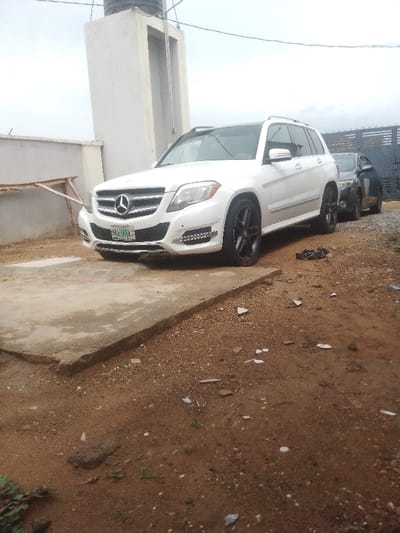 A white Mercedes-Benz ML-Class SUV is parked on a concrete driveway. The vehicle is facing the camera, parked on the right side of the driveway. The front wheels are turned slightly to the right, and the car is parked on a slight incline. The car has black wheels and tires, and the tires are partially obscured by the car's body. There is a metal gate on the right side of the car, partially obscured by the SUV. There is a building in the background on the left side of the car, and some unpaved ground on the right. The ground is covered in dirt, pebbles, and some debris. The location is in Bida, Nigeria, at the coordinates latitude 9.045, longitude 6.003.