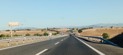 The image shows a highway perspective in the city of Santa Cruz del Retamar, Spain, under a clear blue sky. The highway is divided into multiple lanes, with white lines marking the lanes. A metal guardrail runs along the right side of the road, with a slope at the end. The landscape on either side of the highway consists of dry, rolling hills dotted with sparse trees. In the distance, mountains are visible. Several vehicles, including cars and trucks, are present on the highway, some in motion and some parked. A large billboard structure stands on the left side of the road, with a ladder attached. A power pole is also visible in the distance. The scene appears to be a typical view of a rural highway in Spain during daylight.