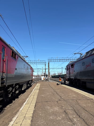 The image depicts a railway station or yard in Kropachyovo, Russia, under a clear, bright blue sky with a single faint contrail. Two large electric locomotives frame the scene, parked on parallel tracks alongside a concrete platform. The locomotive on the left is predominantly red and grey, while the one on the right is grey and red, displaying the "РЖД" (RZD) logo.
The platform, made of cracked concrete with faded yellow tactile paving along its edge, extends into the foreground. Multiple railway tracks converge towards the horizon in the middle ground, featuring extensive overhead electrification lines supported by metal gantries and poles. A small, light-colored utility or signal building stands between the tracks in the mid-distance. Further beyond, green freight cars are visible on additional tracks.
Three individuals are present on the platform. On the right, one person in an orange high-visibility vest stands near the grey and red locomotive, facing towards the tracks. A dark bag rests on the platform nearby. Adjacent to this individual, another person in dark attire stands with their back to the viewer, also near the locomotive. A third person, also in an orange high-visibility vest, is positioned near the small building in the middle distance, facing generally towards the right side of the frame.