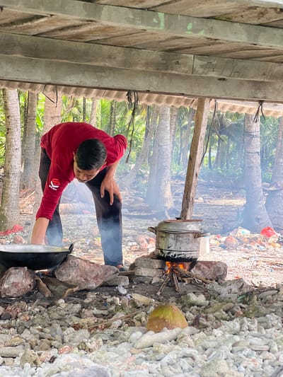 The image shows a person in a red shirt and black pants bending over a cooking setup outdoors. The scene appears to be in a tropical environment, with numerous palm trees in the background and smoke rising from a fire. The cooking area consists of rocks arranged to form a makeshift stove, with a large pot and a steaming pot over the fire. The ground is covered in rocks and debris. This scene is located in Babahie, Indonesia.