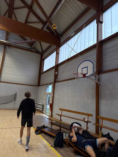 The image captures an indoor sports hall in Sennecey-le-Grand, France. The facility features a light-colored wooden floor with yellow court markings, light grey walls, and a high, sloped ceiling supported by exposed wooden beams and a corrugated metal roof. High on the walls, multiple translucent window panels admit natural light.
Two individuals are present. In the left-center foreground, a male with light-colored, wavy hair stands facing away from the camera, looking upwards. He is dressed in a black long-sleeved shirt, dark shorts, white sneakers, and athletic socks; his left sock has a distinct grey and blue design. Above and to his right, a yellow and blue volleyball is suspended in mid-air near the ceiling structure. On the far left, a portion of a black and white volleyball net is visible.
Along the right side of the hall, several rows of wooden spectator benches are aligned against the wall. A second male, with dark hair, is lying on his back on one of these benches. He wears a dark blue t-shirt with light blue trim and dark blue shorts with light blue accents, holding a small white object near his face. A dark blue backpack, a pair of white sneakers, a green beverage can, and a clear bottle are visible on the bench next to him. Above the benches, a white basketball backboard with a blue outline and a red-rimmed net is mounted on the wall. In the background, towards the left, an exit door with a clear glass panel reveals green vegetation outdoors, and stacks of white poles are stored against the wall.