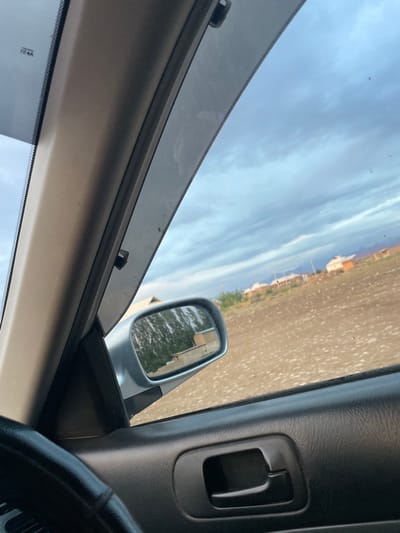 The image is taken from inside a vehicle, looking out through the passenger side window and side mirror. The scene depicts a landscape with a dirt road and buildings in the distance, suggesting a rural or semi-urban setting. The sky is partly cloudy, indicating daytime. The interior of the car is visible, including part of the dashboard, a door panel with a handle, and the steering wheel. The side mirror reflects a glimpse of trees and possibly more buildings. The overall impression is of a journey through a sparsely populated area. There is a small, faint timestamp or serial number visible on the lower windshield, reading "12 B4". The location context provided is Bujum, Kyrgyzstan.