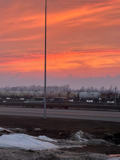 The scene depicts a twilight environment, likely sunrise or sunset, over Melekes, Russia. The sky dominates the upper two-thirds of the image, showcasing vivid orange, red, and pink hues with distinct horizontal cloud formations. In the foreground, patches of dirty snow and melting ice are visible on dark, uneven ground. A multi-lane roadway occupies the lower mid-ground, where two blurred vehicles, a lighter-colored sedan and a darker SUV, are in motion, traveling from left to right. A tall, slender, dark grey pole extends vertically through the center of the frame, originating from the median or roadside. Behind the roadway, a long freight train consisting of numerous cylindrical tank cars, predominantly white or light-colored, is visible. Several tank cars display the "URAL" designation. In the distant background, a dense line of leafless deciduous trees stretches horizontally beneath a hazy, pale purple sky layer.