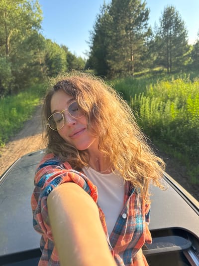 A young woman with wavy, blonde hair and glasses is taking a selfie from an open car sunroof. She is wearing a plaid shirt and a white top, and her eyes are closed with a slight smile. The background shows a dirt road winding through a lush green forest with tall trees and sunlight filtering through the leaves. The scene appears to be in Ol'khovo, Russia, possibly during the summer due to the vegetation and clear sky.