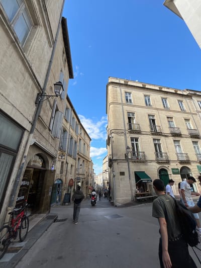 The image depicts a street scene in Montpellier, France, showcasing buildings with a distinctive architectural style. The sky is bright blue with a few clouds. On the right side of the image, people are walking along the sidewalk in front of a building. There appears to be a cafe or restaurant as well. On the left side of the image, a person is walking down the sidewalk. Further down the street, there is a moped or motorcycle. A red bicycle is leaning against the side of a building.