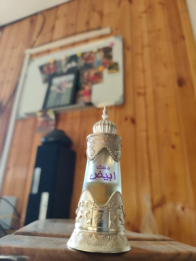 A close-up, low-angle shot features a decorative perfume bottle with ornate silver detailing and a crown-like top. The bottle is clear glass, containing a light-colored liquid. Purple Arabic script is visible on the bottle. The perfume bottle rests on a wooden surface, likely a table, in what appears to be an indoor setting with wood-paneled walls. In the background, a black speaker and a whiteboard covered with photographs are out of focus. The scene is likely within a private residence or small business in Edineț, Moldova, given the available context.