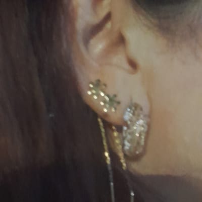 The image shows a close-up of a person's ear with two earrings. The earring on the left is a small, star-shaped stud with multiple points. The earring on the right is larger and appears to be a dangle earring with a cluster of sparkling stones. A thin chain hangs from the right earring. The background is out of focus, with dark hair visible on the left and skin tones on the right. The image does not provide any information about the location in Rudəkənar, Azerbaijan.