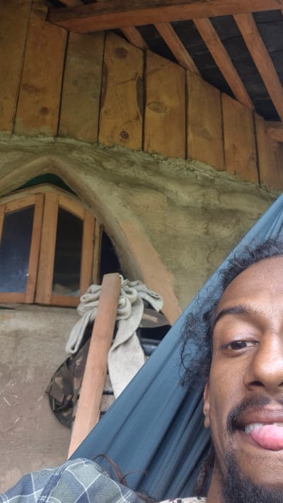 The image shows a close-up, selfie-style shot of a person lying in a hammock. The person's face, particularly their eye, nose, mouth, and beard, are prominently featured in the upper right portion of the frame. They are sticking their tongue out, suggesting a playful or relaxed mood. The background reveals a rustic interior setting. To the left, there are wooden planks forming a wall or structure, with angled wooden beams above. In the center, a rough, textured wall is visible, possibly made of adobe or cob. An arched opening reveals a wooden-framed window with dark panes. A folded cloth or blanket rests on a wooden support beam in front of the opening. The lighting appears natural, suggesting daytime, and there are no immediate clues about the weather. The overall impression is one of a cozy, perhaps handmade living space. The provided location context of "Shila, India" hints at a possible geographical setting, though no specific landmarks or cultural elements are clearly identifiable within the image itself. The person is wearing a plaid shirt on the right side of the image. The composition is slightly tilted, as if taken from a reclining position.