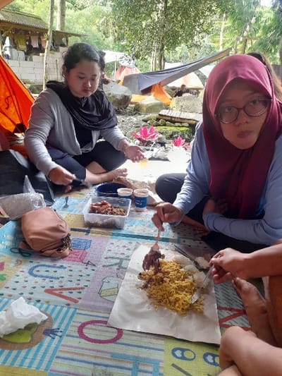 Three people sit on a patterned mat outdoors, seemingly camping. One person wears a maroon hijab and glasses, another a black scarf. Food, including noodles and skewers, is laid out on paper. A clear plastic container of food sits nearby, along with drinks and a brown handbag. Tents are visible in the background, surrounded by trees. The focus is on the group enjoying their meal in a casual, outdoor setting.