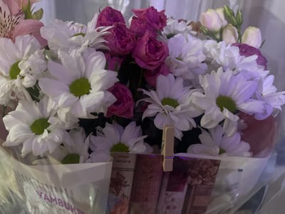 A bouquet of white chrysanthemums and pink roses is presented, wrapped in plastic and newspaper. A wooden clothespin secures a small card or tag to the wrapping. The background appears to be an indoor setting, possibly a room with soft lighting and a textured curtain. The newspaper visible beneath the flowers features text and images, with some of the text appearing to be in Russian. The overall impression is that of a gift or decorative arrangement. The presence of the newspaper in Russian suggests a connection to Russia or a Russian-speaking community, and Shahtinsk, Kazakhstan is a city with a significant Russian-speaking population.