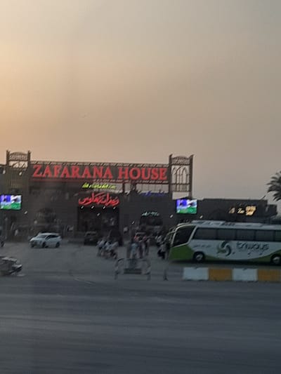 The image shows a building with "ZAFARANA HOUSE" in large red letters on its facade. There is Arabic script below that and electronic billboards on the building. A bus with the word "stridays" in white letters is parked near the building. The setting appears to be an urban area. Based on the location, it's likely the Gharib Cape, Egypt. There are some vehicles on the road, and a palm tree is visible in the background.