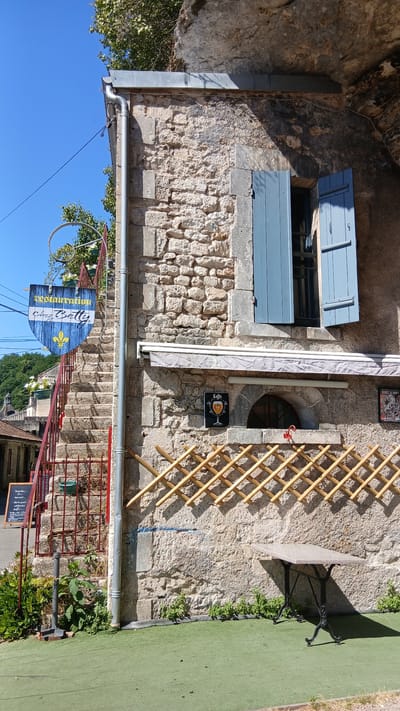 The image shows an outdoor scene of a building facade with stone and stucco walls. A blue-shuttered window is visible on the upper left of the building. To the right of the window, a circular opening is set into the wall. A bamboo trellis stands against the wall, and a small table with a metal base and stone top is positioned next to it. A patch of bright green turf is on the right side of the frame.
A sign for "Restaurant Chez Betty" hangs from a red metal structure, pointing towards the lower right. The sign is blue with yellow lettering and a fleur-de-lis symbol. Below this, a black chalkboard sign is propped up.
There are no people visible in the image. The lighting suggests it is daytime, with distinct shadows cast by the trellis and table, indicating a sunny day. The overall setting appears to be a quaint, possibly historic, establishment in Brantôme en Périgord, France, likely a restaurant or cafe. The stonework and shutters evoke a traditional European architectural style. The presence of the chalkboard and restaurant sign indicates it is an active business.