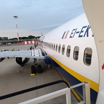 The media file captures an active scene at an airport, likely Orio al Serio, Italy, during dusk or early evening. The primary subject is a Ryanair Boeing 737 aircraft, identifiable by its distinctive white, blue, and yellow livery. An Irish flag emblem is painted near the front of the fuselage, along with the partial aircraft registration "EI-EK".
Passengers are actively boarding or disembarking the aircraft. Several individuals are visible on the front mobile stairs leading to the main cabin door, while others are seen walking across the wing towards a rear exit, suggesting the use of both front and rear stairs for efficiency. Ground staff, some wearing high-visibility vests, are also present on the tarmac near the aircraft and the wing.
Notable details include the aircraft's engine and landing gear, with yellow chocks visible near the wheels. The sky above is a soft, muted blue-grey, indicating the late hours of the day. In the background, airport infrastructure is visible, including a tall red and white striped light pole, parked cars, and distant airport buildings.
Visible text on the inside of the aircraft's door frame reads, "FOG MUST BE ACCOMPANIED WINDOW WHEN SLIDE IS ENGAGED," next to a red arrow pointing downwards, likely a safety instruction for emergency exits. The overall scene depicts a busy, routine moment of air travel at an airport.
