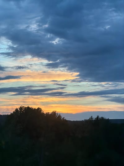 The image presents a view of the sky at dusk, with a portion of what seems like a wooded area visible on the right side. The sky transitions from dark blue clouds on the left to stripes of pale yellow and blue clouds towards the right, close to the silhouette of trees. The light is dimming, as evidenced by the dark silhouettes of trees and the fading colors in the sky. The image does not provide any information regarding the location in the city of Arsk, Russia.