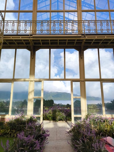 The image shows an interior view of a building in Candikuning, Indonesia, featuring large windows that offer a view of a lake surrounded by mountains. The building has a balcony with an ornate metal railing. Below, the interior space opens to the outside, with the view framed by the building's structure. Purple flowers and other greenery are planted in front of the building. The windows reflect the sky and clouds, creating a sense of depth.