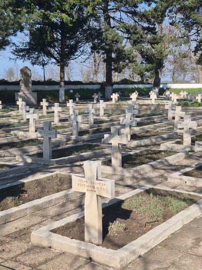 The image shows a cemetery with rows of similar-looking, light-colored stone grave markers. The markers are mostly in the shape of crosses. Trees are visible near the left side of the image, and the sky is visible in the upper left. The markers are surrounded by concrete frames with barren or partly vegetated ground in between. Text is inscribed on many of the markers, appearing to be in Bulgarian. One marker reads "ПОЛК. ЛАЗАР ХР. ВЪРБАНОВ 31 П.П. BAPHA". The image is rotated. The location may be Shumentsi, Bulgaria.