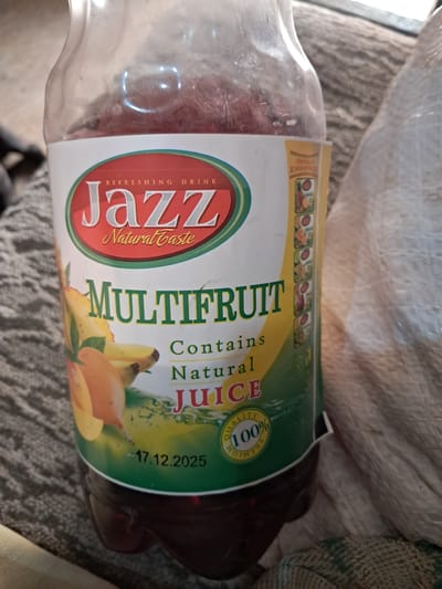 A close-up shot shows a bottle of Jazz brand multifruit juice with a label that reads "Refreshing Drink" and "Natural Taste." The label features images of fruit, including mango and banana. Below the fruit images, text indicates it "Contains Natural Juice." A seal on the label reads "Quality 100% Premium." The expiration date on the bottle is "17.12.2025." The bottle appears to be partially filled with a dark red liquid. The background is out of focus and shows a textured surface, possibly fabric. The image is taken in Chobruchi, Moldova.