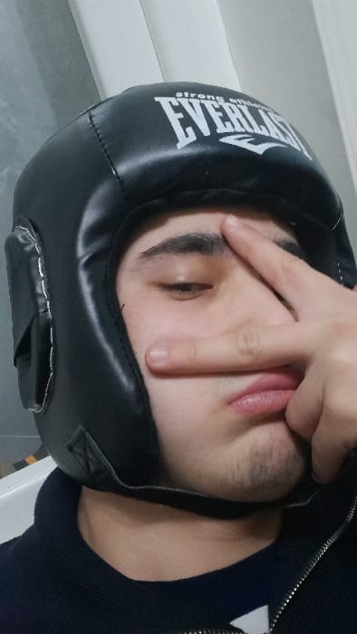 A person wearing a black Everlast boxing headgear is lying down. Their eyes are partially closed, and their hand is raised with two fingers extended in a peace sign near their face. The person has dark hair and a slight stubble. They are wearing a black jacket. The background appears to be a plain white wall or surface.