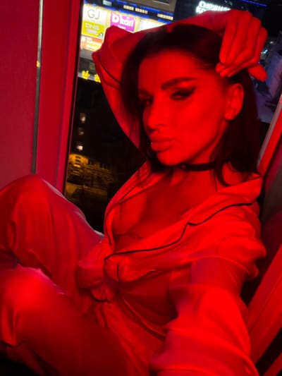 A person is indoors, near a window, bathed in red light. The person is wearing what appears to be a satin pajama set with dark trim. They are touching their hair with one hand. Outside the window are buildings, including one with a sign that reads "DKart" and "DNS", indicating a commercial area. The city is Svkh Voronezhskiy, Russia.