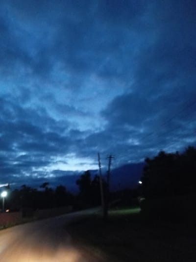 The image depicts a dimly lit, rural scene at dusk or dawn in Adzhimchyghra, Abkhazia. A paved road curves from the foreground into the middle ground, with indistinct, dark shapes of trees and foliage lining its edges. In the distance, a mountain range is visible, silhouetted against a deep blue sky filled with scattered, dark clouds. Several utility poles with visible wires are scattered throughout the landscape, and a single, illuminated lamppost stands on the left side of the road. The lighting suggests low visibility, with the headlights of a vehicle potentially illuminating the road in the foreground. There are no people or discernible activity visible in the image.