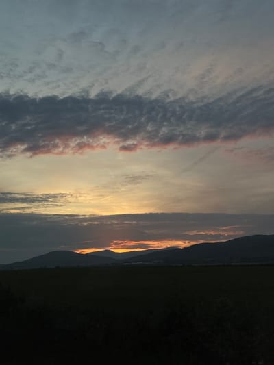 The image displays a captivating sunset or sunrise over a natural landscape, likely in Gavrailovo, Bulgaria. The sky dominates the scene, characterized by a dramatic interplay of light and cloud formations.
Near the horizon, an intense glow of golden yellow and bright orange radiates, indicating the low position of the sun behind a dark, undulating silhouette of mountains or hills. This light illuminates the lower edges of dense cloud banks, creating a luminous band across the sky. Above this, a prominent horizontal stretch of clouds takes on soft pink and reddish hues, contrasting with the lighter, pastel gray and blue tones of the surrounding sky. Towards the upper and left portions of the frame, the clouds appear more diffused and scattered, ranging from light gray to darker, textured patches, suggesting varied altitudes and densities.
The landscape below the horizon is cast into deep shadow, appearing as a dark, almost black, expanse. The contours of the mountains are discernible against the brighter sky, but the foreground consists of an undifferentiated mass of land, likely fields or distant forests, entirely unlit by the setting or rising sun.
There are no people, animals, or man-made structures visible. The scene captures a tranquil moment in nature, emphasizing the serene beauty and rich colors of the sky at dawn or dusk. No visible text is present in the media file.