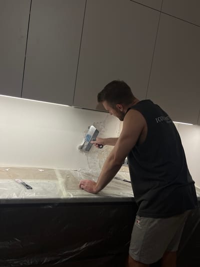 A man is using a trowel to apply plaster to a wall. He is wearing a black tank top and gray shorts. The wall is white and appears to be partially plastered. The floor is covered in plastic sheeting. The location in the city of Svkh Voronezhskiy, Russia is not identifiable from the image.