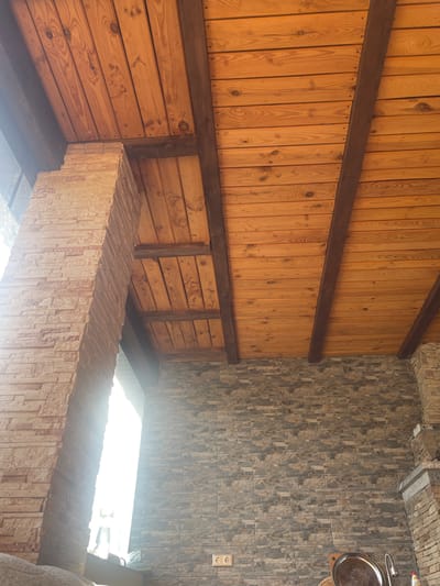 This image, located in Petrivka, Ukraine, captures a view of a covered outdoor or interior space, primarily focusing on its architectural elements from a low-angle perspective looking upwards. The dominant feature is the ceiling, constructed from numerous light-to-medium brown wooden planks laid horizontally, secured by visible nails. These planks are supported by several thicker, dark brown exposed wooden beams that run perpendicularly across the ceiling.
On the left side of the frame, a large, vertical structural pillar is visible, constructed from light-toned, stacked stone veneer with a rough, irregular texture. This pillar is heavily backlit by intense, blinding sunlight streaming from an unseen opening or window on the far left, creating a strong glare and obscuring details beyond.
The wall on the right and across the background is also clad in stacked stone veneer, featuring a mixture of gray and brown irregular-shaped stones. Towards the lower left of this wall, two white dual electrical outlets are affixed. In the bottom right corner, a glimpse of a reflective, circular silver tray with embossed detailing is visible, placed on what appears to be a counter or surface, adjacent to a metal faucet or tap, suggesting a utility or kitchen area. No people are present in the scene. The overall environment conveys a rustic or natural architectural style under bright daylight.