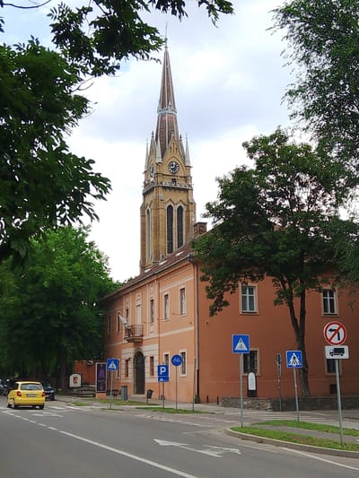 A yellow car drives down a street in front of a large, light-orange building with a tall, gothic-style tower. Trees with green leaves partially obscure the view of the building and tower. The tower has a clock face and a pointed spire. There are several street signs visible, including a pedestrian crossing sign, a parking sign, a bicycle lane sign, and a no-left-turn sign with a truck symbol. The weather appears to be overcast, and the time of day is daytime. The scene is urban, with a road, sidewalks, and a building that looks like a church or a municipal building. The overall mood is calm and ordinary.