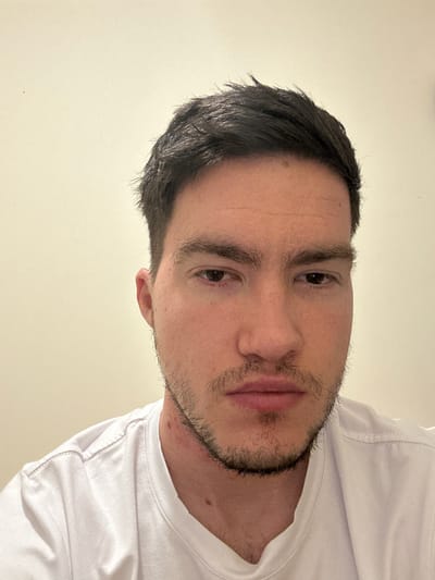 A close-up headshot of a man with short, dark hair and a stubble beard. He is wearing a white t-shirt. The background is a plain, light-colored wall. It is impossible to locate the scene in the city of Forest of Dean, United Kingdom based on the image.