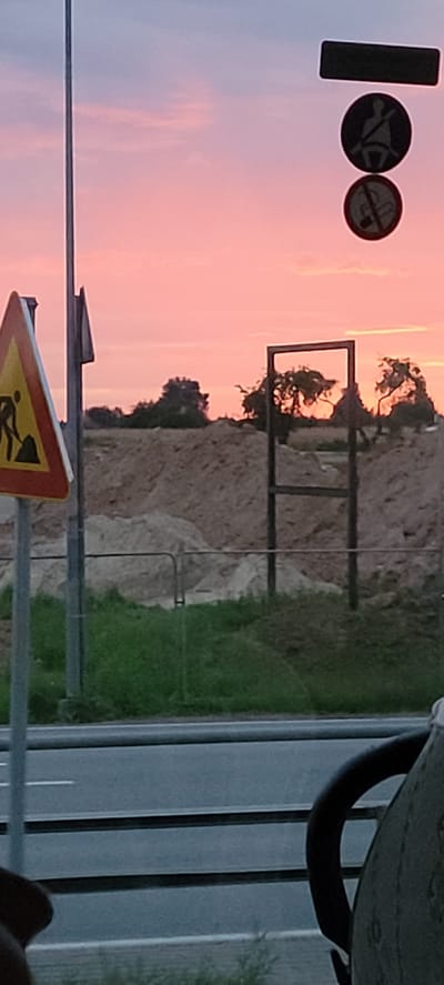 The image shows a construction site with a pink sunset sky in the background. A traffic sign indicating work is in progress is visible on the left. Several traffic signs are also present, including one for seatbelt usage and another for no smoking. A metal frame structure stands in the middle of the scene, with trees visible behind it. The foreground shows a road and a grassy area. The image appears to be taken in Iecava, Latvia.