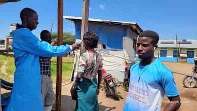 The image depicts an outdoor scene in Tchere, Democratic Republic of the Congo, during a bright, sunny day with a clear blue sky. The central activity involves a person in a light blue disposable protective gown and a black face mask, who is smiling and using a white infrared thermometer to take the temperature of a woman. The woman, seen from behind, wears a floral-patterned long-sleeve top and a dark green skirt, holding a black handbag.

Behind the health worker, another man wearing a plaid shirt and a dark face mask is partially visible. In the foreground to the right, a young man with earbuds looks off-camera to his left with a neutral expression. He is wearing a blue and white t-shirt that displays text.

The setting appears to be a semi-rural or community area. Structures made of wood and blue tarpaulin are visible behind the individuals, suggesting temporary dwellings or service points. Further in the background, a blue and white building with several solar panels on its roof can be seen. The ground is unpaved and dusty. Two motorcycles are parked near the structures, and a large black water tank is visible on the left.

Visible text on the t-shirt of the man in the foreground reads "SIS WATER", followed by "minerale naturelle pure", "ozonisé, filtré par barrière", and "eau minerale souterraine traitée aux uv", indicating a water brand. The name "Joseph Kisuki" is watermarked on the bottom right corner of the image.