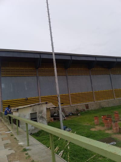 The image depicts a section of Obafemi Awolowo University in Ile-Ife, Nigeria. A large building with grey corrugated metal walls and yellow louvered openings dominates the background. A tall, grey metal pole with ladder-like protrusions runs vertically in the frame. In the foreground, a concrete walkway with a green metal railing is visible. Two individuals are sitting on the railing near a small, dilapidated structure with a window. The ground slopes away from the building, with green vegetation interspersed with items such as rusted barrels and other debris. The sky appears overcast.