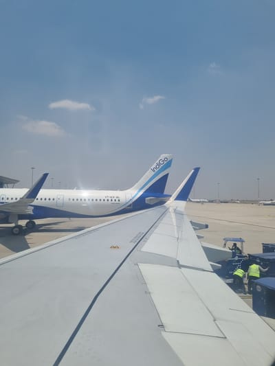 The image captures a view from inside an airplane, looking out over the wing. Another Indigo airplane is visible on the tarmac, with its tail and Indigo logo clearly displayed. Airport ground personnel in bright yellow safety vests are positioned around a luggage conveyor. The sky is blue with some clouds. The tail of the plane says it is VT-ICR. The tarmac is made of cement.