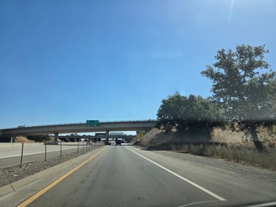 This media file captures a view from the driver's perspective on a highway in Sacramento, California. The scene is set on a clear, sunny day with a bright blue sky, suggesting it is daytime. The highway has multiple lanes, with a concrete barrier and chain-link fence lining the left side.
A prominent overpass stretches across the highway, with traffic visible on it. A green road sign on the overpass indicates "SOUTH I-80 TRUXEL ROAD 1 MILE". To the right of the highway, a dry, grassy embankment is topped with trees, casting shadows on the ground, hinting at a warm climate. A single tree with a broad canopy stands near the embankment. The overall impression is one of travel on a typical sunny day in a semi-arid or Mediterranean climate. No people are clearly visible. The activity is vehicular travel on a highway.