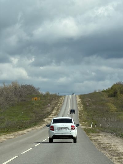 A roadside view in Michurinsky, Russia shows a white Lada automobile parked on the right shoulder of a two-lane road. A dark vehicle is visible further ahead on the same road. The road is flanked by grassy terrain with sparse vegetation on both sides. Overcast skies dominate the upper half of the frame.