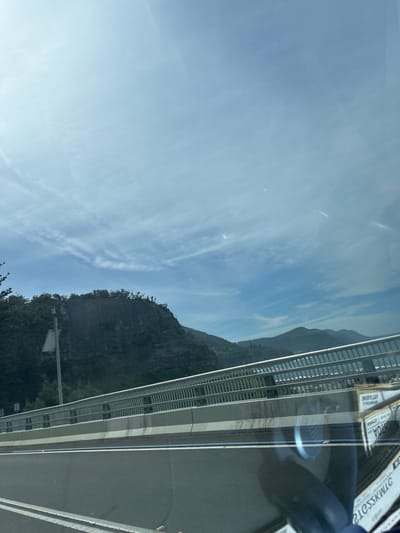 The image depicts an exterior view captured from within a vehicle, showcasing a section of the Sea Cliff Bridge area in Coalcliff, Australia. The scene is set under a light blue sky with scattered, wispy white clouds, indicating daylight conditions. The bottom portion of the frame shows the vehicle's dark dashboard or interior, displaying reflections of light and partially legible, inverted text.
In the midground, a multi-lane roadway is visible, marked with white and blue lines. This road is bordered by a concrete barrier and a robust metal railing system, characterized by multiple horizontal bars and vertical posts. Beyond the railing, to the left, a prominent, sheer dark grey rock cliff face rises steeply, its summit densely covered with green foliage. A light-colored utility pole, fitted with a rectangular panel, stands adjacent to the cliff on the far left. To the right of the cliff and beyond the bridge railing, a calm body of water, identified as the Pacific Ocean, is visible. Further in the distance, a series of verdant hills and mountain ranges recede towards the horizon. No individuals, specific actions, or interactions are discernible within the depicted environment.