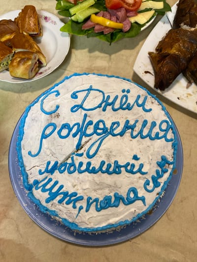 An overhead shot depicts a celebratory scene with food items arranged on a light brown surface. A round cake is centered and prominently features white frosting with a blue border and blue writing in what appears to be Russian Cyrillic. A slice has been cut from the cake. To the left of the cake, there is a white plate with sliced pastries or pies. Another white plate presents a salad featuring lettuce, cucumbers, tomatoes, sliced sausage, and cheese. In the upper left corner, a white rectangular plate holds smoked fish. The scene likely portrays a celebration, possibly a birthday, given the presence of a cake with celebratory writing. Based on the Russian Cyrillic text on the cake, it could be located in a Russian-speaking region, and given the task constraints, it can be identified as Тушна, Russia.