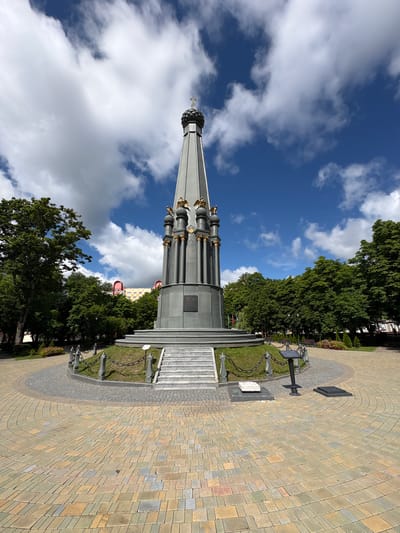 This media file shows a monument in Polatsk, Belarus. The monument is tall and conical, with ornate golden decorations and a cross at the very top. It is surrounded by a paved walkway made of colorful bricks that form a curved pattern. A chain-link fence encircles a grassy area around the monument. In the background, lush green trees line the path, and a few buildings are visible beyond the trees. The sky is a vibrant blue with scattered white clouds. The overall scene suggests a bright, sunny day. The image is oriented vertically, with the monument pointing upwards towards the sky. There is no visible text in the image.