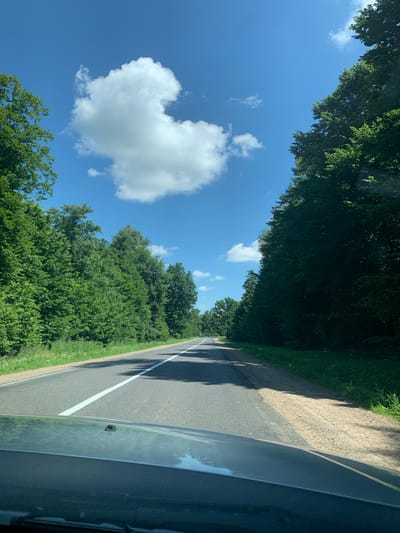 The image captures a view from a vehicle traveling along a paved road bordered by lush green trees on the left and a gravel shoulder with more greenery on the right. The road has a white line marking the center. The sky is a vibrant blue with scattered white, fluffy clouds. The sun appears to be shining, casting shadows of the trees onto the road. The overall scene suggests a pleasant, daytime drive through a rural or semi-rural area. The time of day is likely midday or early afternoon, given the bright sunlight and clear sky. There are no people or visible text in the image. The foliage suggests it is summertime.